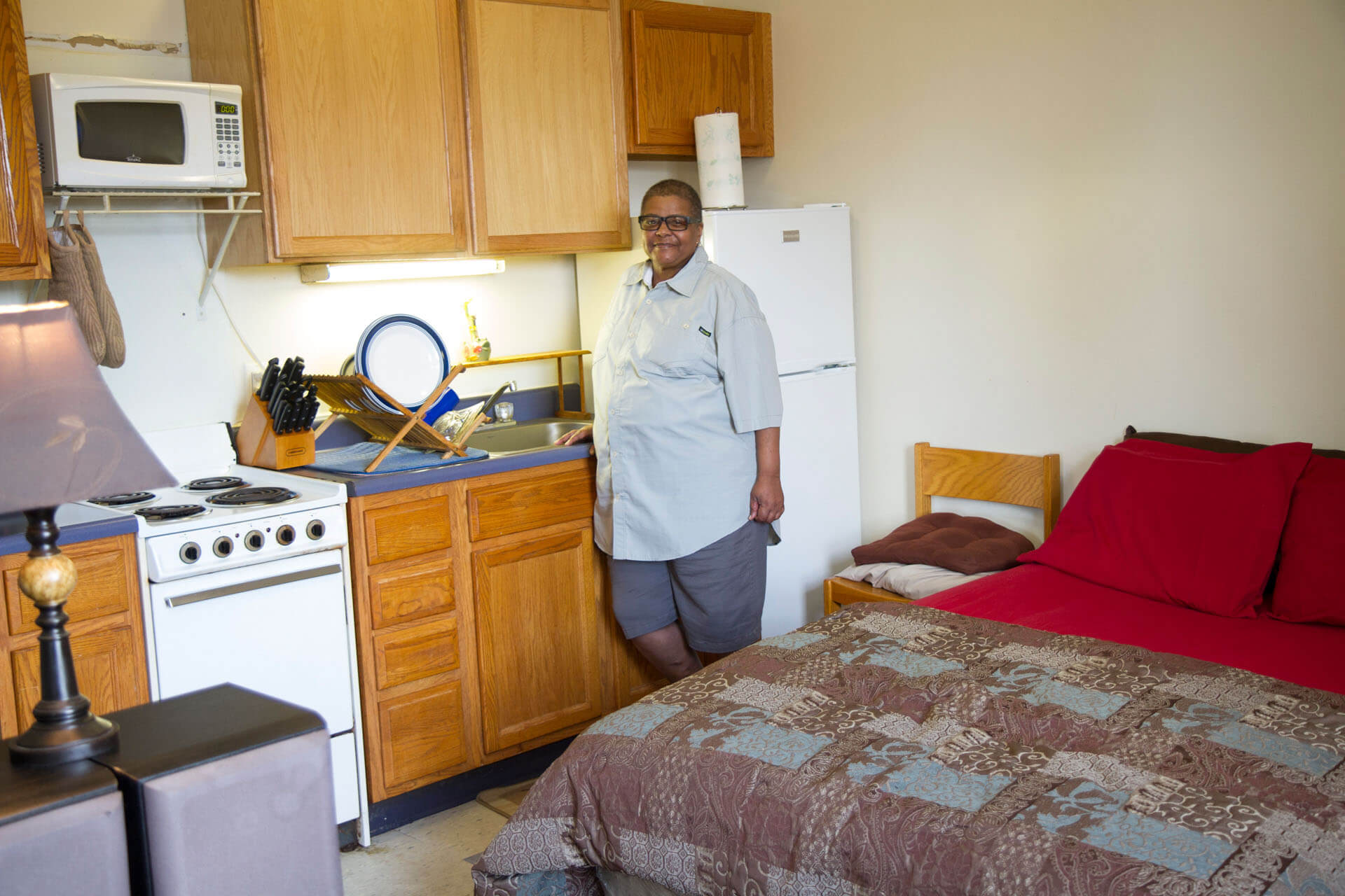 A female tenant in Deborah's Place housing stands in her apartment. She is smiling and leaning on the kitchen counter in her studio apartment.