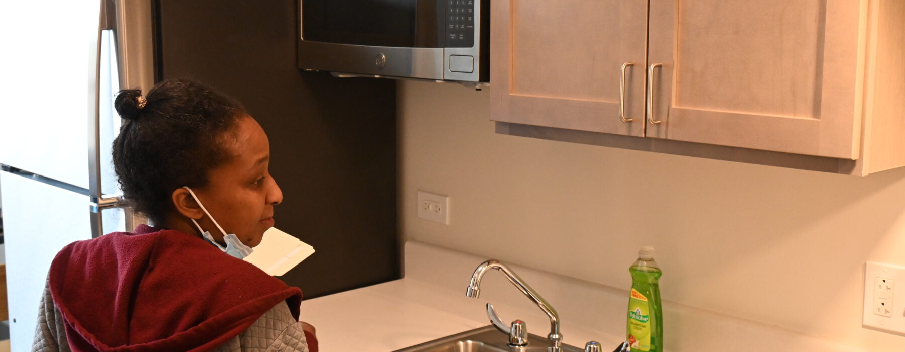 A young woman, Deborah's Place participant, stands in the kitchen of her new apartment. On the coutner are a full cooking utensil set and cleaning supplies provided for her to start life in her new permanent supportive housing.