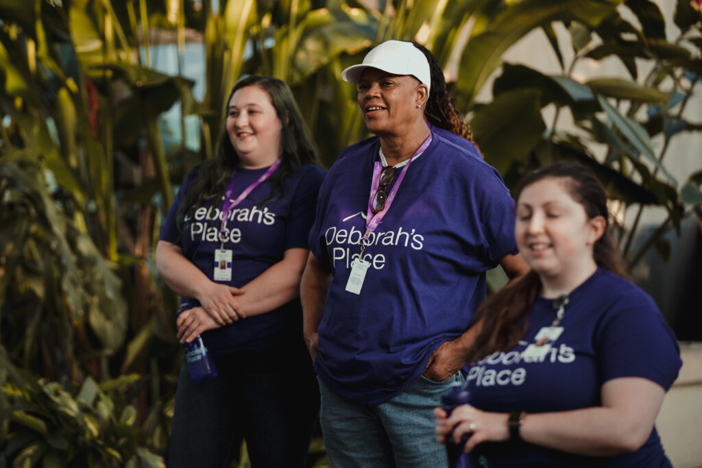 3 Deborah's Place employees in purple shirts and lanyards stand in the Garfield Park Conservatory.