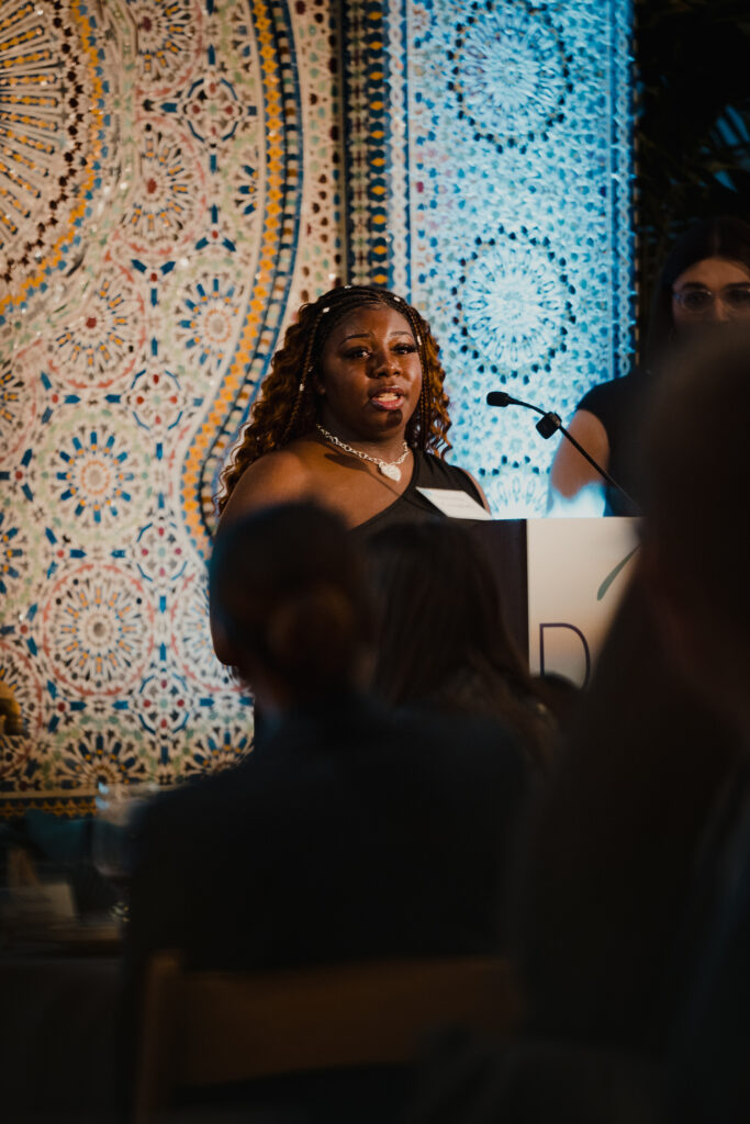 Darnella Campbell, a Black woman with long hair, stands at a podium and speaks