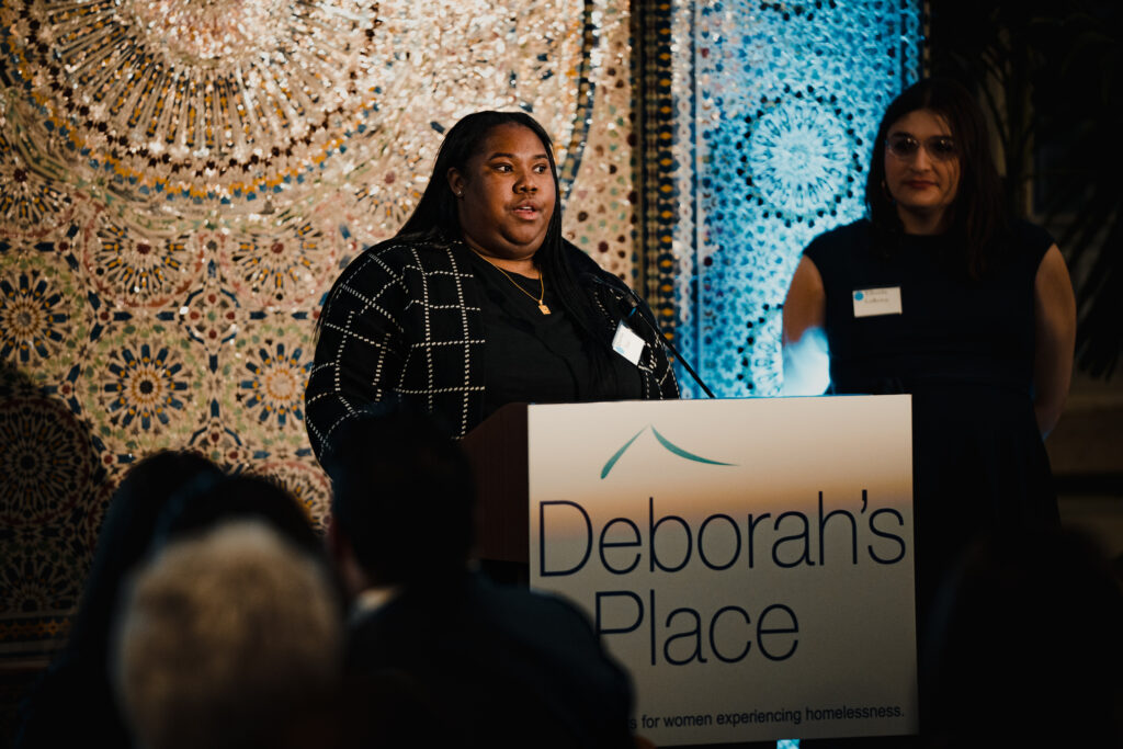 Quashay Tartt, a Black woman in a black checked shirt, stands at a podium and speaks.