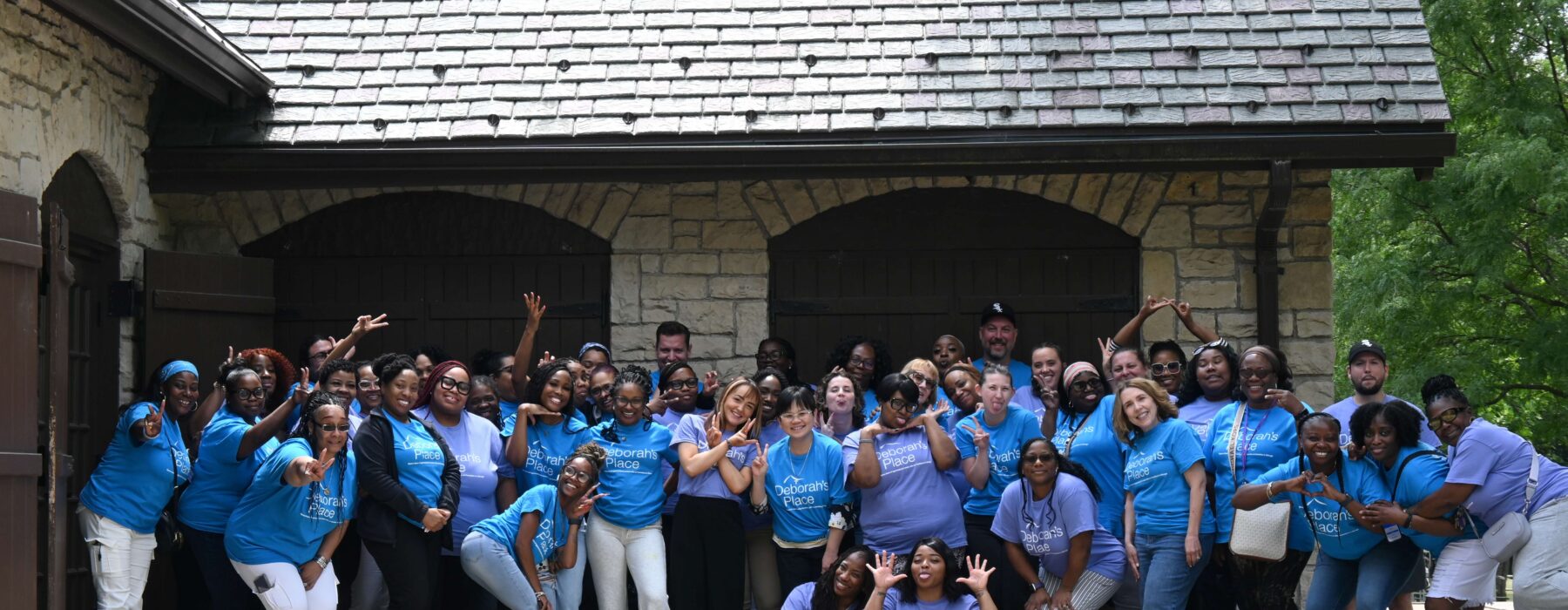 Photo shows almost all full-time Deborah's Place staff, about 50 people, standing in front of a stone building, wearing matching purple and blue t-shirts and smiling.